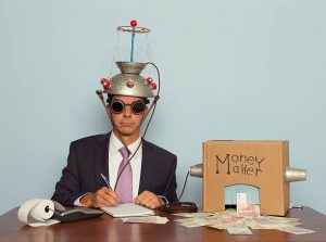 A businessman sitting at a desk records the amount of British Pound Sterling his machine makes from the ideas in his head. He is dressed in a suit and purple tie, glasses, and a mind reading helmet on his head. Bling.